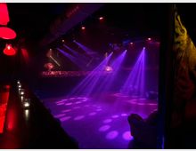 Ein Innenraum einer Disco oder eines Nachtclubs mit einer Tanzfläche, die von violettem und rotem Bühnenlicht beleuchtet wird. Im Vordergrund ist eine lange Bar mit Kerzen und Hockern zu sehen, während sich im Hintergrund eine erhöhte DJ-Kabine mit Blumen befindet.