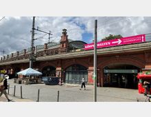 Backsteinbahnhof "Hackescher Markt" in Berlin mit bogenförmigen Eingängen, Schienen und Oberleitungen auf einem erhöhten Bahnsteig. Im Vordergrund befinden sich Gehwege, ein Verkaufsstand, Gehende sowie ein Schild mit einem Hinweis auf den Fernsehturm Alexanderplatz.