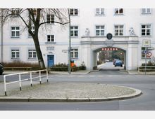 Wohnhaus in heller Putzfassade mit blau gerahmten Fenstern und einer blauen Tür, flankiert von einem großen Torbogen mit Skulpturen und Beschilderung "Schillerhof". Im Vordergrund eine Straße mit Verkehrsinseln und Gehwegbelag aus Kopfsteinpflaster.