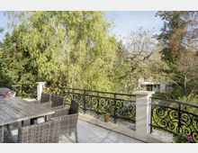 Terrasse mit Steinboden und einem Holztisch umgeben von geflochtenen Stühlen, begrenzt durch ein schmiedeeisernes Geländer mit verschnörkeltem Muster. Im Hintergrund üppige Bäume und ein Wohnhaus inmitten eines Gartens.