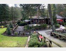 Holzgebäude in einem Waldumfeld, umgeben von Wegen mit Holzgeländern, Grünflächen und kleinen Nadelbäumen. Im Hintergrund ist ein Schild mit der Aufschrift „Café“ und ein rotes Vordach zu sehen.