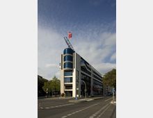 Modernes Bürogebäude mit heller Fassade und großen Glasfenstern an einer Straßenecke. Auf dem Dach befinden sich eine rote Fahne und ein schräg aufgerichtetes Metallgitter, während die Umgebung von Bäumen und Straßenlaternen geprägt ist.