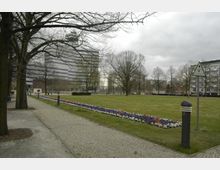 Ein Park mit einer großen Rasenfläche, gesäumt von blühenden Blumenbeeten in Weiß, Rot und Blau. Im Hintergrund stehen kahle Bäume, ein modernes Bürogebäude und mehrstöckige Wohnhäuser.