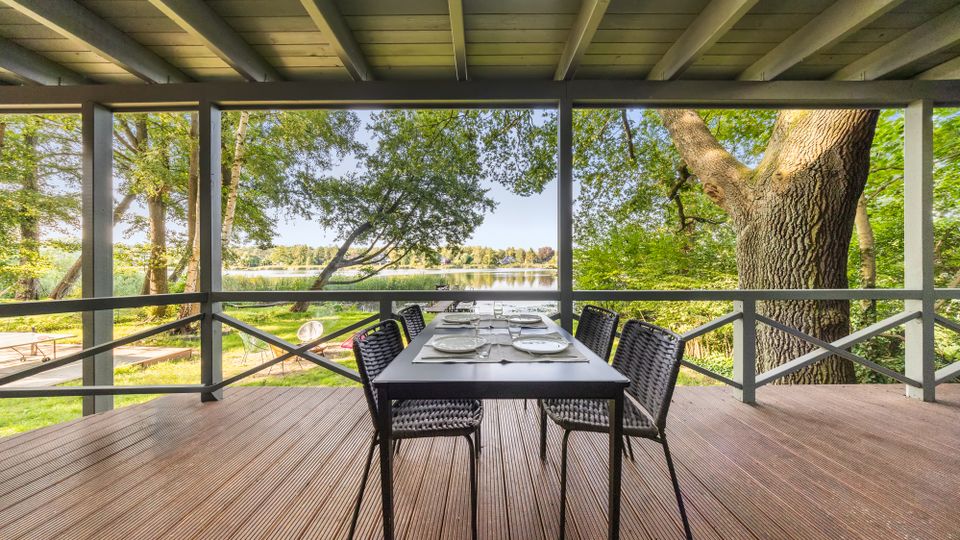 Überdachte Holzterrasse mit Tisch und vier Stühlen, eingerahmt von Geländer und Deckenbalken. Blick durch Bäume auf einen See und eine grüne Uferlandschaft, mit großem Baumstamm am rechten Rand.