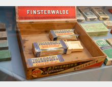 Holzkiste mit Seifenprodukten der Marke "Thierack's Kernseife Gut Wetter", darunter verpackte und ausgepackte Seifenstücke, umgeben von weiteren historischen Seifenverpackungen. Der Deckel der Kiste trägt den Schriftzug "Finsterwalde".
