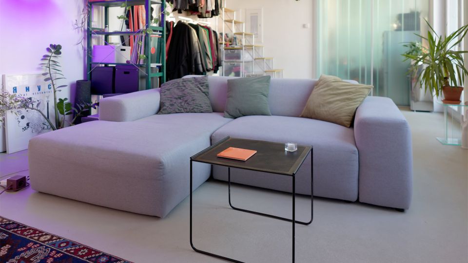 Modernes Wohnzimmer mit großem hellgrauem Ecksofa, kleinem schwarzem Metall-Couchtisch und mehreren Zimmerpflanzen. Im Hintergrund stehen offene Regale und eine Kleiderstange, daneben eine Treppe sowie eine Milchglas-Schiebetür.