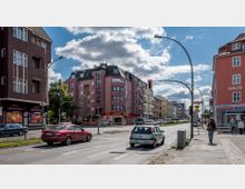 Straßenszene mit mehrstöckigen Wohn- und Geschäftshäusern, überwiegend in Pastellfarben gehalten. Im Vordergrund eine befahrene Kreuzung mit Autos, Fußgängern und einer Ampel, umgeben von Gewerbeflächen wie einer Apotheke und einem Spielcenter.