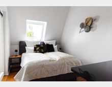 Helles Schlafzimmer mit schräger Decke, einem Doppelbett mit weißer Bettdecke und Kissen in Schwarz und Gold. Neben dem Bett steht ein kleiner Nachttisch mit einer Lampe, und ein schräges Fenster lässt Tageslicht herein; an der Wand hängt eine dekorative Metallkunstinstallation.