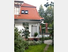 Bild eines Einfamilienhauses mit weißer Fassade, rotem Ziegeldach und Dachfenstern. Im Vordergrund ein gepflegter Garten mit grünem Rasen, Blumen und einem Metallbogen mit Rosengewächsen; daneben ein gepflasterter Weg und eine mit Klinkern verzierte Mauer.