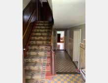 Treppenhaus in einem älteren Gebäude mit einem holzverkleideten Geländer, einem mit gemustertem Teppich bedeckten Treppenaufgang und einem Boden aus gelb-grauen Fliesen. Der Flur führt zu mehreren Türen und einem Raum mit einem Fenster im Hintergrund.