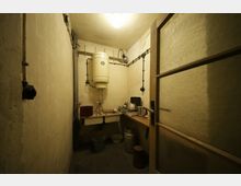 Kleiner, spärlich beleuchteter Raum mit einem Warmwasserboiler an der Wand, einer Doppelspüle sowie einer Arbeitsplatte mit Wasserkocher und Küchengeräten. Der Raum hat beige Wände, sichtbare Rohrleitungen, und einen Steinboden; Eimer und Behälter stehen verstreut.