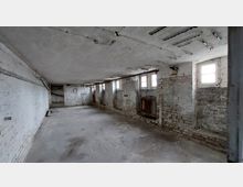 Ein leerer, verlassener Kellerraum mit weiß gestrichenen Ziegelwänden, mehreren kleinen Fenstern an der rechten Wand und freiliegenden Rohren an der Decke. Auf der linken Seite ist eine Türöffnung sichtbar, und an der rechten Wand befindet sich ein montierter Heizkörper.