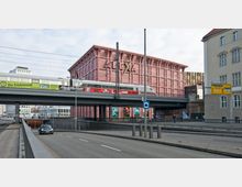 Eine Straße mit einer Brücke im Vordergrund, auf der ein Zug fährt. Im Hintergrund steht das Einkaufszentrum „Alexa“ mit seiner auffälligen rosa Fassade, daneben befinden sich weitere Gebäude, darunter ein älteres, beiges Haus.