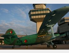 Ein grünes, zweiflügeliges Flugzeug mit Tarnmuster steht auf einem Flughafenvorfeld, im Hintergrund ein Kontrollturm mit moderner Architektur und blauen Glasfronten. Der Himmel zeigt leichte Bewölkung.