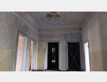 Ein verlassener Innenraum mit stark verblassten Wänden und abblätternder Tapete, drei Türen in einem schlichten Rahmen und einer alten Deckenlampe. Im Hintergrund sind dunkle Gänge und teils verbaute Türöffnungen sichtbar.