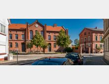 Straßenszene mit zwei historischen Backsteingebäuden; das Gebäude links hat große Rundbogenfenster, ein Uhrturm und ein rotes Ziegeldach, rechts steht ein ähnliches Gebäude mit Schriftzug "Bauverein Wittenberge". Im Vordergrund sind geparkte Autos, Bäume und ein Gehweg sichtbar.