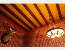 Innenraum mit gelber Decke und sichtbaren Holzbalken, roten Wänden mit gold-orangenen Mustertapeten, einer dekorativen Kronleuchterlampe und einem Hirschkopf als Wanddekor.