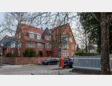 Backsteinbau mit roten Ziegeldächern und mehreren Dachgauben an einer Straßenecke, umgeben von Bäumen und einem niedrigen Backsteinzaun. Im Vordergrund Straßenschilder der Goßlerstraße und Ehrenbergstraße sowie geparkte Autos und ein weißer Holzzaun.