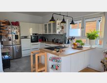 Helle, moderne Wohnküche mit weißen Einbauschränken, Edelstahl-Kühlschrank und einer großen Kücheninsel mit Holzarbeitsplatte und integriertem Kochfeld. Große Fenster mit Blick auf eine Holzverkleidung außen, darüber drei schwarze Pendelleuchten; auf der Insel stehen Schalen, Pflanzen und Küchenutensilien.