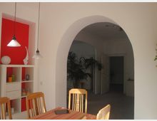 Esszimmer mit einer hellen Einrichtung, einem Holztisch und Stühlen. Im Hintergrund ein rotes Wandregal mit dekorativen Elementen, ein runder Türbogen führt zu einem angrenzenden Raum mit Türen und einer Palme.