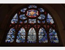 Großes gotisches Kirchenfenster mit kunstvoll gestalteten Buntglasscheiben in Blau-, Rot- und Weißtönen, die eine Rosette und spitzbogige Muster formen. Die Fenster befinden sich in einem steinernen Gewölberahmen.