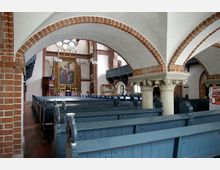 Innenraum einer Kirche mit Ziegelmauerwerk und Rundbögen, die von verzierten Säulen getragen werden. Mehrere Reihen grauer Holzbänke führen zu einem kunstvoll gestalteten Altar mit einem Gemälde, im Hintergrund farbige Glasfenster.