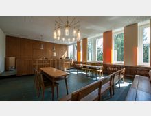 Heller Raum mit großen Fenstern, hölzerner Wandverkleidung und Sitzmöbeln aus hellbraunem Holz. Ein Kronleuchter mit Glaslampen hängt von der Decke, der Raum ist minimalistisch eingerichtet und scheint für Versammlungen oder Arbeit genutzt zu werden.