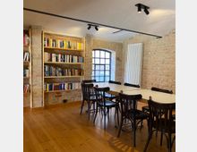 Ein kleiner, gemütlicher Raum mit freiliegenden Ziegelwänden, einem großen Tisch mit mehreren schwarzen Stühlen und einem Bücherregal an der Wand. Ein Fenster mit schwarzen Gitterstäben lässt Tageslicht herein, und der Holzboden verstärkt die warme Atmosphäre.