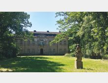 Historisches Herrenhaus mit symmetrischer Fassade und Satteldach, umgeben von Bäumen und einer großen Grünfläche. Im Vordergrund steht eine steinerne Skulptur auf einem Sockel.