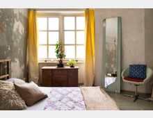 Ein schlicht eingerichtetes Schlafzimmer mit einer verwitterten Wand, einem großen Fenster mit gelben Vorhängen, einem Holzbett und einer Kommode. Ein schmaler Spiegel lehnt an der Wand, daneben steht ein gepolsterter Stuhl mit blauem Kissen.