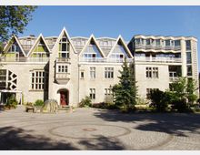 Steinernes Gebäude mit modernem und historischem Architekturstil, charakterisiert durch spitze Dachgauben und rechteckige Fenster. Im Vordergrund ein gepflasterter Platz mit einem Brunnen sowie Bäumen und Pflanzen.