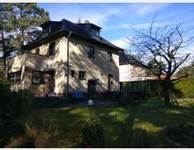 Zweistöckiges Wohnhaus mit heller Fassade und dunkelgrauem Dach in einem Garten mit Grasfläche, Bäumen und Sträuchern. Im Vordergrund steht ein kahler Baum, rechts ein Zaun, und hinter dem Haus befindet sich eine Terrasse mit Außentreppe aus Metall.