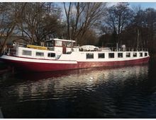 Ein langes, weiß-rot gestrichenes Boot liegt an einem Ufer vor einer Kulisse aus kahlen Bäumen. Das Boot hat rechteckige Fenster entlang der Seiten und ein erhöhtes Steuerhaus.