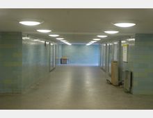 Unterführung mit blaugrünen Kacheln an den Wänden, mehreren an der Decke befestigten runden Lampen und gläsernen Schaufenstern auf der rechten Seite. Am Ende des Gangs steht ein Mülleimer, und der Boden besteht aus hellen Fliesen.