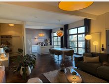 Open-plan modern living and dining room with a white kitchen island, pendant lights, and dark wood herringbone flooring. Large grid-style windows and glass doors with dark curtains open to a balcony, with a sofa and low coffee table in the foreground.