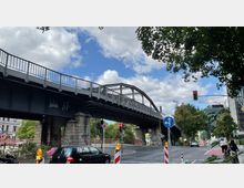 Eine städtische Straßenkreuzung mit einer Stahlbrücke im Bogen-Design, die von Steinsäulen getragen wird. Im Vordergrund sind eine Ampel, ein blauer Verkehrskreiselhinweis und mehrere Fahrzeuge sowie Radfahrer zu sehen, umgeben von Bäumen und moderner Architektur im Hintergrund.