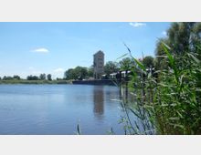 Flusslandschaft mit einem hohen, schmalen Gebäude am Ufer, das von Bäumen umgeben ist; im Vordergrund sind Schilfpflanzen und ruhiges Wasser sichtbar.