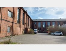 Backsteingebäude eines ehemaligen Industriekomplexes mit großen Sprossenfenstern, umgeben von einem gepflasterten Hof, auf dem mehrere geparkte Fahrzeuge stehen. Im Vordergrund befinden sich Büsche und niedrige Bäume.