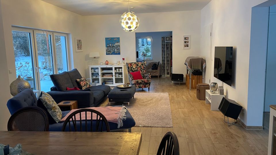 Helles, modernes Wohnzimmer mit großen Fenstern zur verschneiten Außenansicht, grauem Ecksofa mit Ottoman, Teppich und Couchtisch sowie einer auffälligen Kugel-Deckenleuchte. Rechts hängen ein wandmontierter Fernseher und ein kleines Lowboard; im Hintergrund führen Durchgänge in weitere Räume mit Sesseln und Regalen.