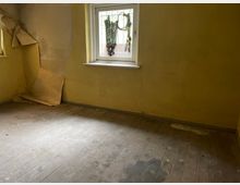 Ein leerer, vernachlässigter Raum mit gelben Wänden, einem teilweise abblätternden Wandbelag in der Ecke und einem kleinen Fenster mit Gitter. Der Boden besteht aus abgenutzten Holzdielen, und der Raum wirkt unrenoviert.