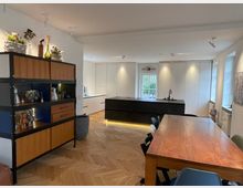 Moderner Wohnraum mit offener Küche und Essbereich. Im Vordergrund steht ein großer Holztisch mit Stühlen, im Hintergrund eine Kücheninsel mit dunkler Oberfläche und weißen Einbauschränken; links ein Regal mit Dekorationen, der Raum hat lichtdurchflutete Fenster und Fischgrät-Holzboden.