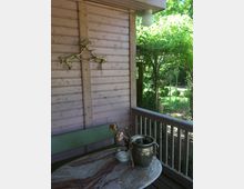 Holzveranda mit Blick ins Grüne, an der eine pastellfarbene Holzfassade mit einem Hirschgeweih dekoriert ist. Auf einem Marmortisch stehen ein metallener Blumentopf mit einer vertrockneten Pflanze und ein kleines Gefäß; eine Holzbank und ein Geländer ergänzen die Einrichtung.