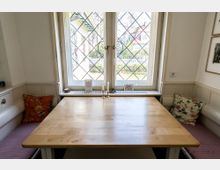 Gemütliche Essecke mit einem Holztisch, umgeben von gepolsterten Sitzbänken mit bunten Zierkissen. Im Hintergrund ein großes Sprossenfenster mit Blick auf einen Garten und Häuser im Freien.