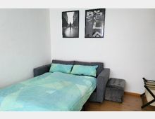 Wohnzimmer mit ausgezogenem Schlafsofa, das mit mintgrüner Bettwäsche bedeckt ist, darauf zwei Kopfkissen. An der weißen Wand hängen zwei schwarz-weiße Bilder, daneben steht ein grauer Polsterhocker. Der Boden ist aus Holz.