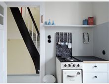 Kleine Küche mit einem weißen Elektroherd, schwarzem Spritzschutz und hängenden Küchenutensilien. Rechts daneben befinden sich eine Arbeitsfläche, ein Regal mit Dekoartikeln und links eine offene Tür, die den Blick auf eine Treppe mit schwarzem Geländer ermöglicht.