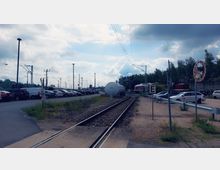 Bahngelände mit einem Gleis, das durch eine offene Fläche führt, gesäumt von parkenden Autos auf beiden Seiten. Im Hintergrund sind Oberleitungen, ein großer weißer Tank und abgestellte Züge zu sehen, umgeben von Bäumen und Wolken am Himmel.