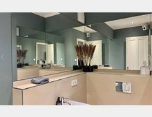 Modernes Badezimmer mit grauen Wänden, großem Wandspiegel und beiger Wandverkleidung. In der Ecke steht dekoratives Pampasgras in einer Vase, und auf dem weißen Waschbecken liegen Handseife und ein gefaltetes Handtuch.