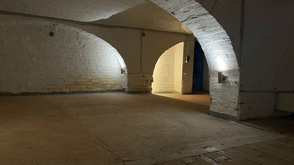 Gewölbter Kellerraum mit weiß gestrichenen Backsteinwänden und großen Rundbögen, beleuchtet durch wandmontierte Lampen. Der Raum hat einen glatten Betonboden und führt durch einen Bogen in einen helleren Nebenbereich.