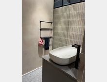 Modernes Badezimmer mit einer hellen, gefliesten Wand, einem ovalen weißen Waschbecken auf einer grauen Arbeitsplatte und einem schwarzen Wasserhahn. An der gegenüberliegenden Wand befindet sich ein Handtuchhalter mit einem pinken und einem dunklen Handtuch.
