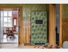 Alt-Text: Innenraum eines Hauses mit einer grünen, verzierten Kachelheizung im Vordergrund. Im angrenzenden Raum stehen ein Holztisch und Stühle auf einem schwarz-weiß gemusterten Boden, vor einer rosa Wand mit einem vergoldeten Spiegel. Die Wände im Vordergrund sind holzvertäfelt und grün tapeziert.